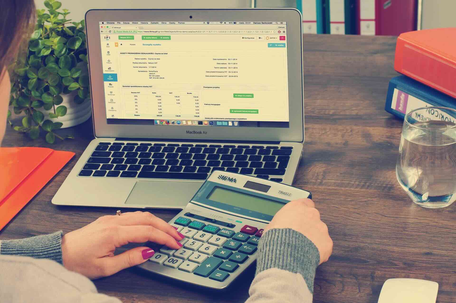 Woman's profile and hands holding large calculator, in front of laptop screen display a spreadsheet - WebGrit
