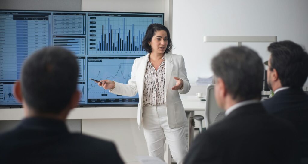 Young woman standing in front of large monitor, pointing to charts, making BPO presentation to businessmen - WebGrit