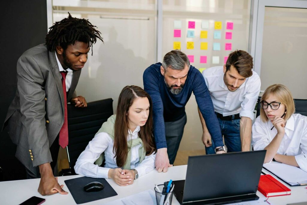 5 people staring at laptop in conference room, one middle-aged and rest young, post-it notes on whiteboard in background - WebGrit