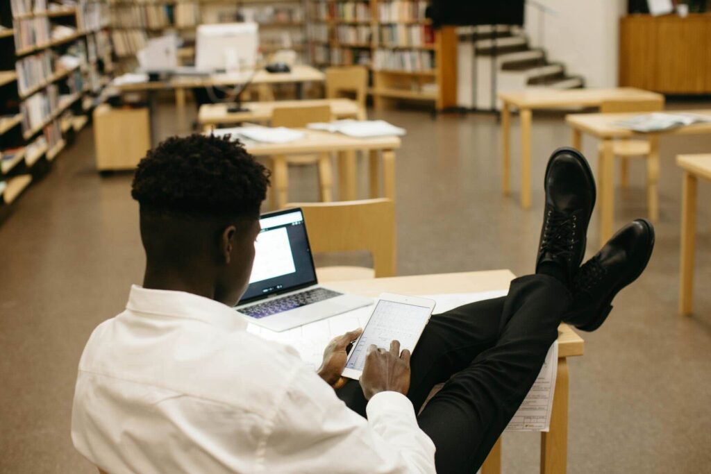 Back view of young black man with legs propped on desk, looking at tablet, print library in background, laptop open on desk - WebGrit